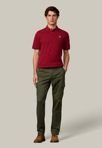 Bordová polo tričko s golierom, zelené cargo nohavice a hnedé topánky. Tričko má logo na ľavej hrudi a kontrastné šitie.