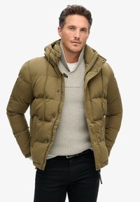 Chaqueta de plumas verde oliva con capucha desmontable, cremallera frontal y botones a presión. Suéter de punto gris texturizado por debajo, jeans negros.