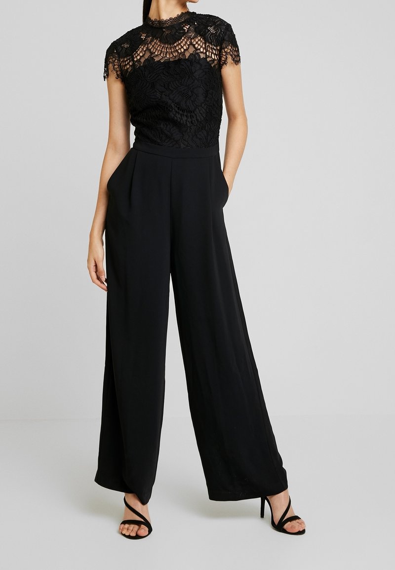 Femme portant un pantalon large noir et un haut en dentelle noire à manches courtes, debout les mains dans les poches avec des talons noirs à brides.