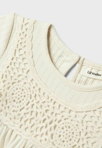 Haut crème avec un tissu côtelé texturé et des détails en crochet élaborés le long de l'encolure. L'étiquette indique "Lil' Atelier".