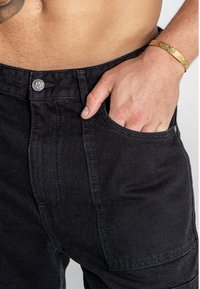 Zwarte denim broek met een voorzak, metalen knoopsluiting en stiksel details. De stof heeft een gladde textuur.