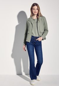 Světle zelená ribbed bunda s předními kapsami, doplněná o ladící košili a tmavě modré zvonové džíny. Bílá teniska dokončuje celý outfit.