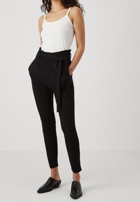 Osoba stojí oblečená v bílém tílku a černých kalhotách s vysokým pasem, doplněná opaskem a lesklými černými muly na neutrálním pozadí.