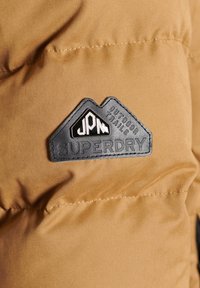 Détail d'une doudoune marron avec un patch en caoutchouc noir portant un texte blanc. Le patch a une forme triangulaire et une couture visible.