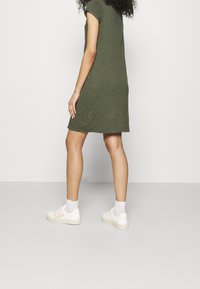 Robe courte à manches courtes de couleur olive, longueur genoux, en tissu texturé. Portée avec des baskets blanches et des chaussettes de cheville, debout sur un sol gris.