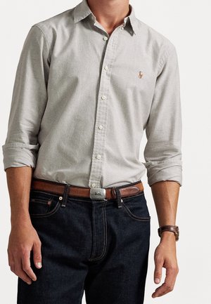 Mand iført lysegrå skjorte med knapper og opkrogede ærmer, mørke jeans, brun læderbælte og brun armbåndsur, stående mod hvid baggrund.