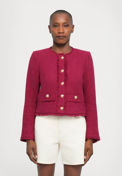 Fuchsia, gestructureerde jas met rafelige randen, gouden knopen en twee voorkruiden. Verkorte stijl en lange mouwen. Gekleed met witte shorts.