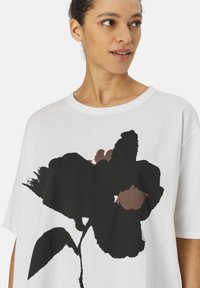 Witte katoenen t-shirt met een grote zwarte bloemenprint en bruine accenten. Losse fit en ronde halslijn. Zachte textuur.