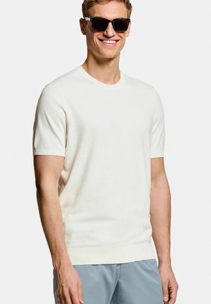 Jonge man die glimlacht, draagt zwarte zonnebril, een wit shirt met korte mouwen en lichtblauwe broek, staande tegen een effen achtergrond.