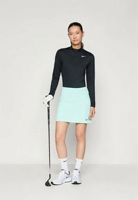 Melna garenās piedurknes sporta kreklā, piparmētru zaļa golfa svārku, balta golfa cimds, baltas kurpes, melni Nike logo akcenti, stāv ar golfa nūju.