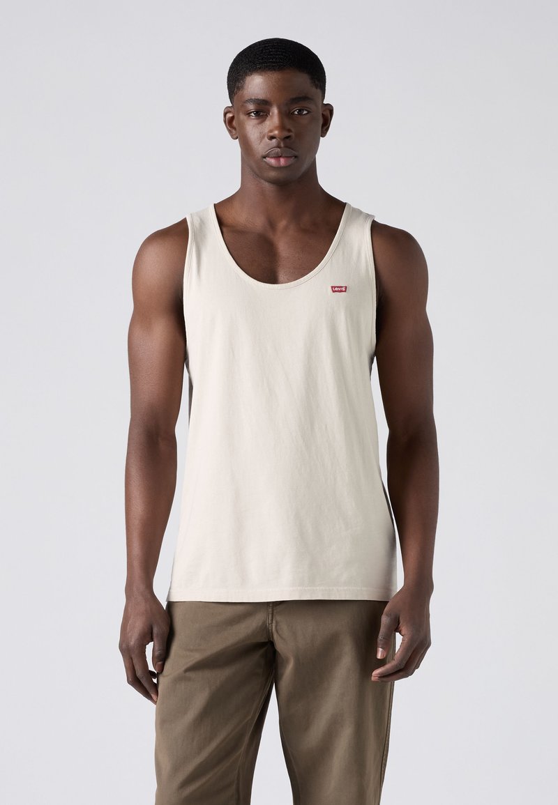 Jeune homme portant un débardeur beige sans manches et un pantalon marron, debout devant un fond gris clair uni.