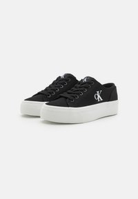 Pantofi sport negri din pânză cu talpă din cauciuc alb. Caracteristicile includ un design cu șireturi și un mic logo alb pe lateral. Siluetă plată, stil minimalist.