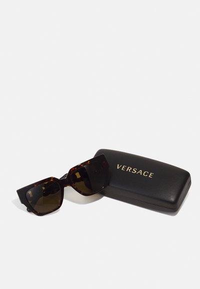 Mustade ja kilpkonnakuju suurte päikeseprillide tumedate läätsedega, koos musta nahata kandikuga, millel on kuldsed "Versace" brändimärgid.