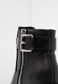 Bottine en cuir noir avec une fermeture éclair latérale, une sangle ajustable avec une boucle argentée, et une texture lisse avec des détails de couture subtils.