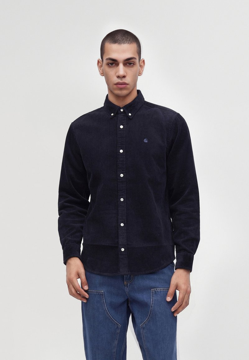 Jeune homme portant une chemise en velours côtelé noire boutonnée et un jean bleu, debout devant un fond clair uni.