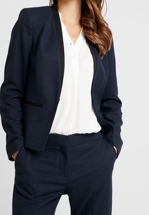 Vrouw in een marineblauwe blazer en broek met een witte blouse, handen in de zakken, tegen een effen lichte achtergrond.