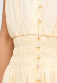 Robe jaune pâle avec une surface texturée, caractérisée par une taille froncée et une fermeture boutonnée à l'avant avec des boutons blancs.