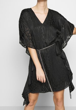 Personne portant une courte robe noire texturée avec des manches larges et une ceinture chaîne argentée, debout devant un fond clair uni.