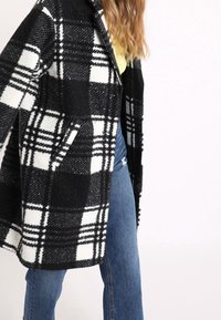 Cappotto a quadri bianco e nero con una consistenza morbida, apertura frontale, e tasche laterali, abbinato a jeans azzurri chiari.
