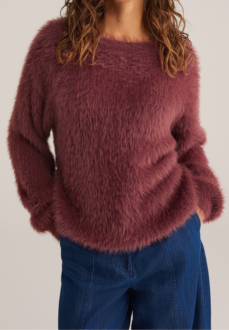 Femme portant un pull duveteux à manches longues de couleur mauve foncé, les mains dans les poches d’un pantalon en jean bleu taille haute.