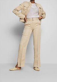 Beige jasje met twee borstzakken, gecombineerd met bijpassende broek met zijsplits. Draagt bijpassende geweven instappers.