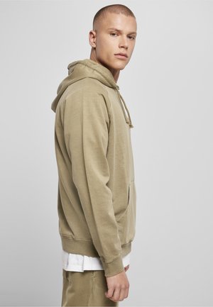 Fiatal férfi rövid hajjal, bézs kapucnis pulóvert és nadrágot viselve, oldalról áll egy sima, világosszürke háttér előtt.