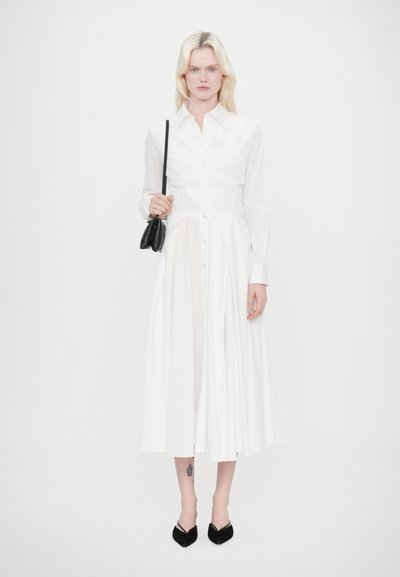 Vestido blanco de botón, hecho de un tejido ligero, con un corpiño ajustado y una falda fluida. Lleva unos tacones negros de punta y un pequeño bolso negro.