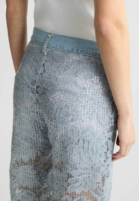 Pantaloni in pizzo blu chiaro con motivo floreale, abbinati a una vita in denim e dettagli in paillettes. Presentano un design aderente e una finitura testurizzata.