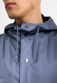 Veste à capuche bleu clair avec un col montant, des boutons-pression en métal, des accents à cordon de serrage et un tissu lisse et résistant à l'eau.