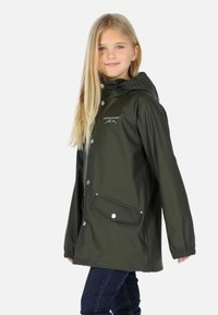 Jeune fille portant un imperméable vert foncé à capuche, debout de profil, souriante, et portant un jean bleu foncé sur un fond uni.