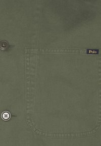 Detailní záběr na zelenou látkovou košili s přední kapsou, na které je malá nášivka značky Polo a viditelné knoflíky po straně.