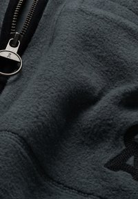 Tessuto in pile grigio con una consistenza morbida, caratterizzato da una cerniera inclinata e un dettaglio del logo nero. Il materiale sembra spesso e caldo.