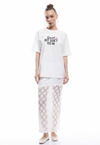 Witte oversized t-shirt met de tekst "Sweet but don't test me" graphic, gecombineerd met een witte kanten rok met bloemvormige uitsparingen en lichtgekleurde schoenen.