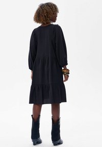 Vrouw met krullend haar in een zwarte gelaagde jurk, zwarte cowboylaarzen tot halverwege de kuiten en twee grote armbanden, met de rug naar ons gekeerd.