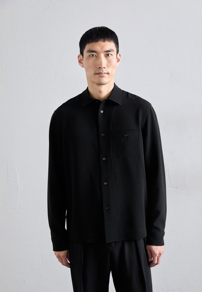 Camisa negra de botones hecha de tela suave, con un bolsillo en el pecho izquierdo y cuello clásico, combinada con pantalones negros.