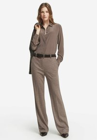 Brun långärmad blus med knäppning framtill, bärs med ljusbruna, vida byxor och en texturerad mörk sjal. Svart bälte och skor.