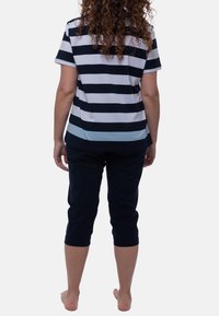 Nainen, jolla on kiharat hiukset, seisoo paljain jaloin. Hänellä on päällään musta-valkoraidallinen lyhythihainen paita ja mustat puolipitkät housut, ja hän katsoo poispäin.