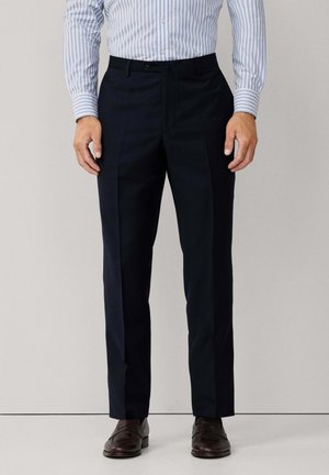 Man draagt zwarte op maat gemaakte broek, een blauw-wit gestreept overhemd en bruine leren loafers, staand tegen een lichte muur.
