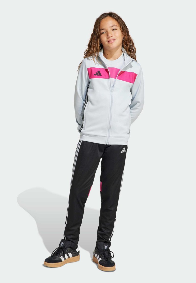 Persona joven de pie con las manos detrás de la espalda, vistiendo una chaqueta con cremallera blanca y rosa, pantalones deportivos negros y zapatillas Adidas negras.