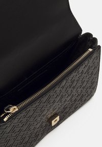 Schwarze Lederhandtasche mit geprägtem geometrischen Muster, Reißverschluss und einem goldfarbenen quadratischen Verschluss. Das Innere ist mit schwarzem Stoff ausgekleidet.