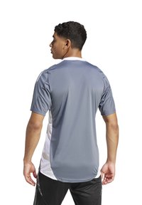 Maglia sportiva grigia e bianca con maniche corte, scollo a giro e texture liscia, caratterizzata da pannelli laterali e accenti in contrasto.