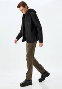 Svart softshell-jacka med huva, kombinerad med olivgröna byxor och svarta stövlar. Slät textur, praktisk design och funktionella detaljer synliga.