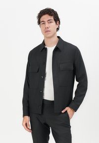 Black button-up jacket with two front pockets, long sleeves, and a structured collar. Worn over a white t-shirt, featuring a smooth texture.