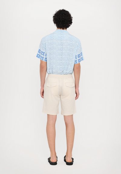 Cămașă cu mânecă scurtă, de culoare albastru deschis, cu un model floral geometric și pantaloni scurți de culoare crem. Sandalele completează look-ul casual.