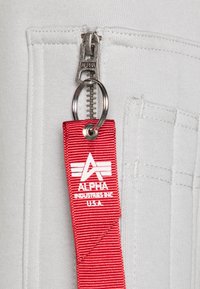 Sivkasta tkanina z zadrgo in srebrno zadrgo. Rdeča oznaka z belim besedilom vsebuje "ALPHA INDUSTRIES INC. U.S.A." in grb.