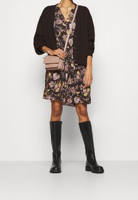 Schwarze Strickjacke über einem blumigen braunen Kleid, kombiniert mit knielangen schwarzen Stiefeln und einer kleinen rosa Umhängetasche. Auffällige Knöpfe an der Strickjacke.