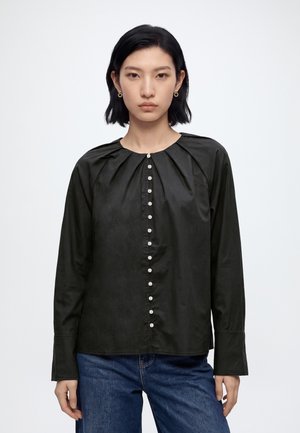 Femme aux cheveux noirs coupés au carré, portant un chemisier noir boutonné à manches longues et un jean bleu sur un fond clair uni.