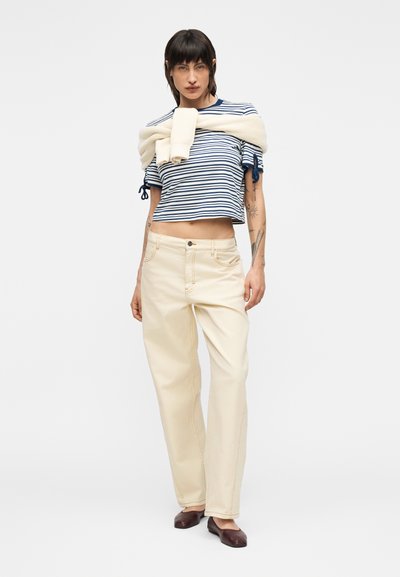 Γυναίκα που φοράει μπλε ναυτικό και λευκή ριγέ κοντή μπλούζα, κρεμ παντελόνι φαρδιάς γραμμής, καφέ μπαλαρίνες και ένα κρεμ πουλόβερ που είναι περασμένο στους ώμους, στέκεται σε λευκό φόντο.