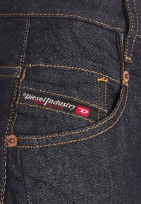 Mörka denimjeans med kontrasterande orange sömmar, med en märkesetikett i svart och rött på framlommen och metallaccenter.