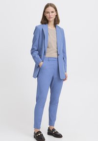 Completo su misura blu chiaro con blazer strutturato e pantaloni slim-fit, abbinato a un top beige e mocassini neri con dettagli dorati.